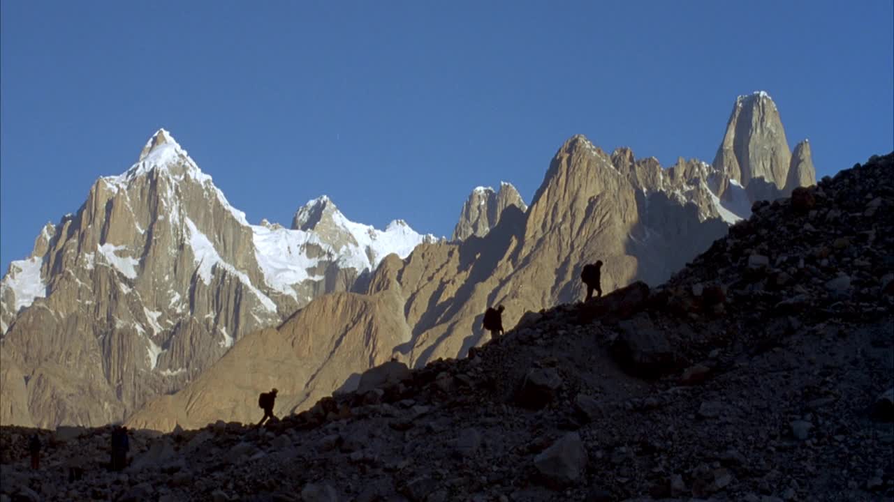 几个登山者沿着岩石斜坡攀登的广角。登山者在山的阴影里。在背景中看到金字塔形状的山峰。山峰被雪覆盖着。在两座山峰之间可以看到陡峭的冰川。视频素材