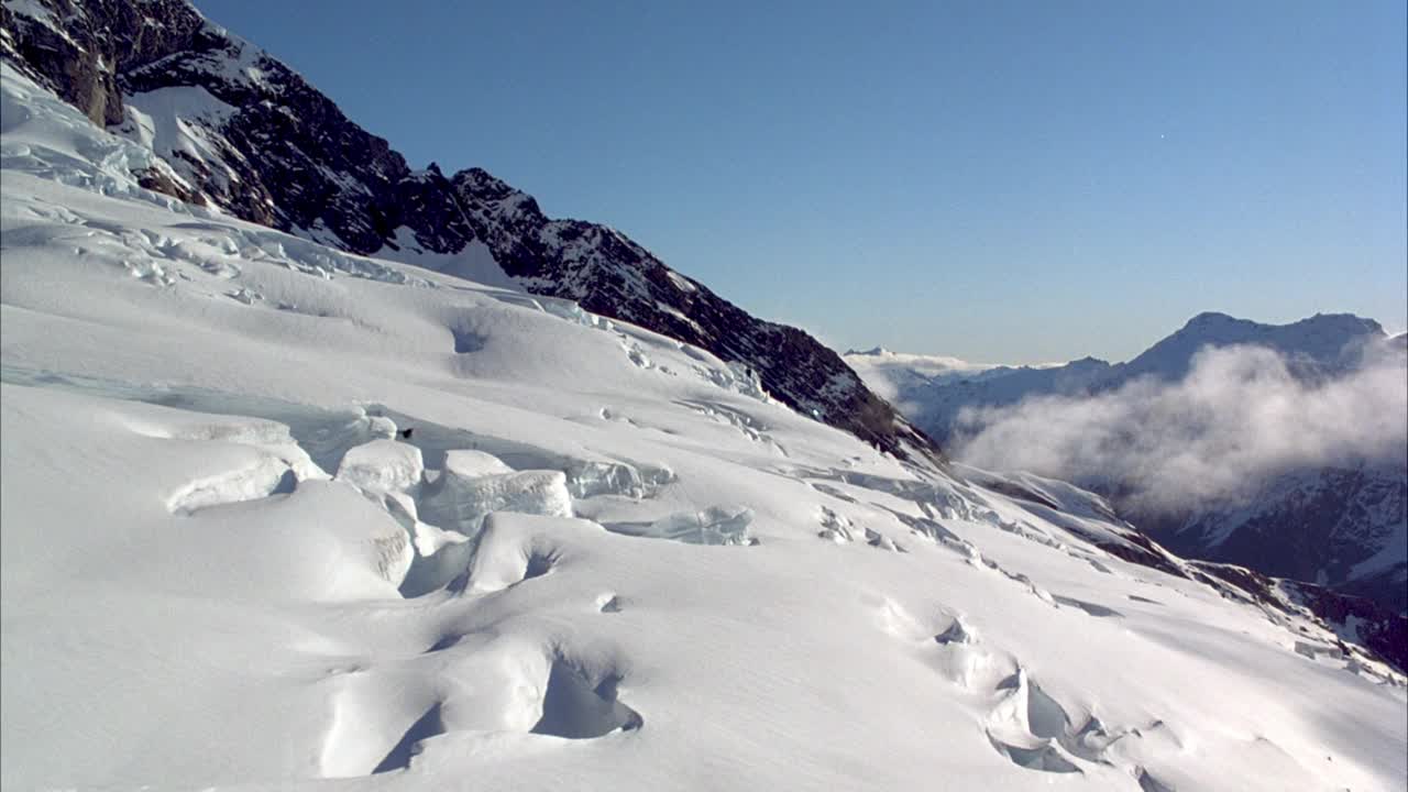 在积雪覆盖的山坡或冰川上的空中飞行。看到冰川上的低雾或云。偶尔可以看到右上方的直升机叶片。在背景中可以看到雪山和清澈的蓝天。视频素材