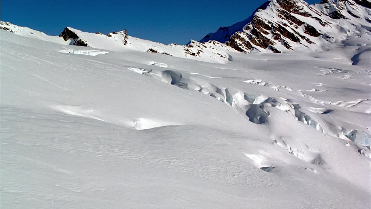 在积雪覆盖的山坡或冰川上的空中飞行。在背景中可以看到雪山和清澈的蓝天。视频素材