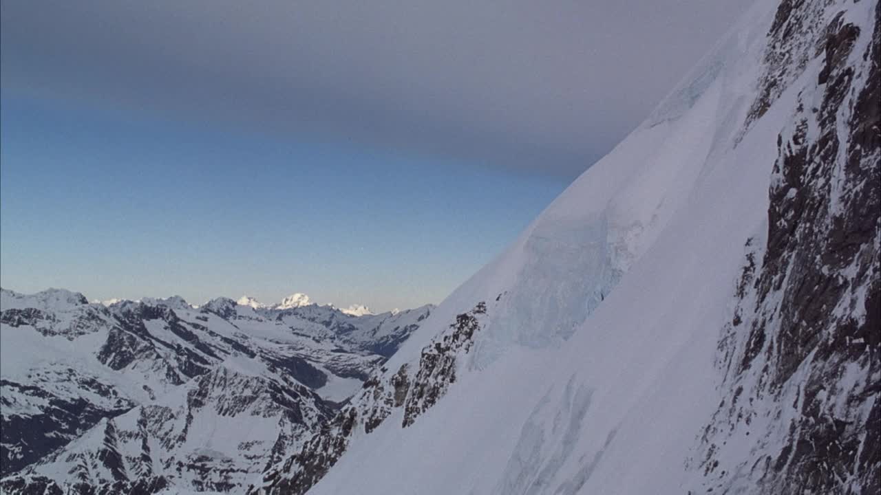 沿被雪覆盖的山顶的山坡横向架空。放大山坡上冰川的形成。在较低的背景下，可以看到高坡周围的低云和蓝天和雪山。视频素材