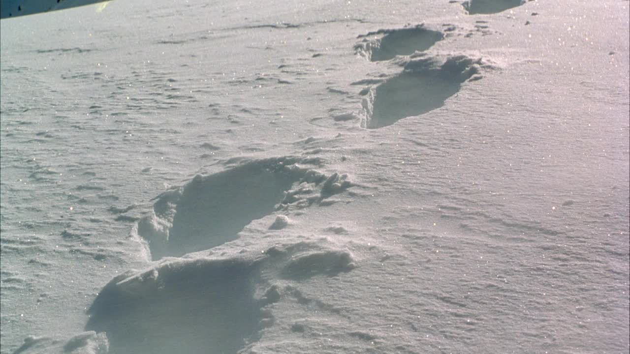 中等角度的脚印在深雪斜坡上。看雪从左到右吹上斜坡。看雪在阳光下闪闪发光。向上扒，露出两位登山者在远处尽头的脚印。看到攀登者沿着积雪覆盖的山脊行走。年代视频素材