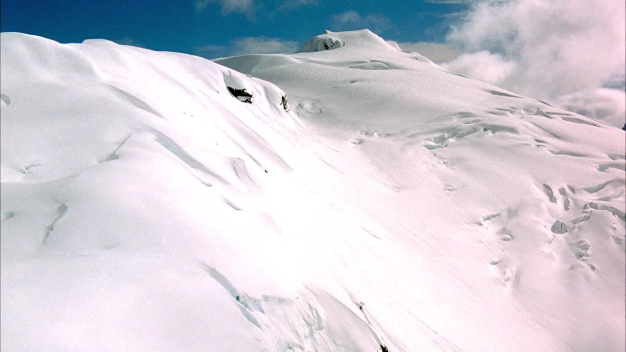 在积雪覆盖的山坡上的空中飞行。可能是山坡上的冰川。当pov沿着山脊线飞行时，看到山顶上低垂的积云。在背景中可以看到积云和蓝天。视频素材