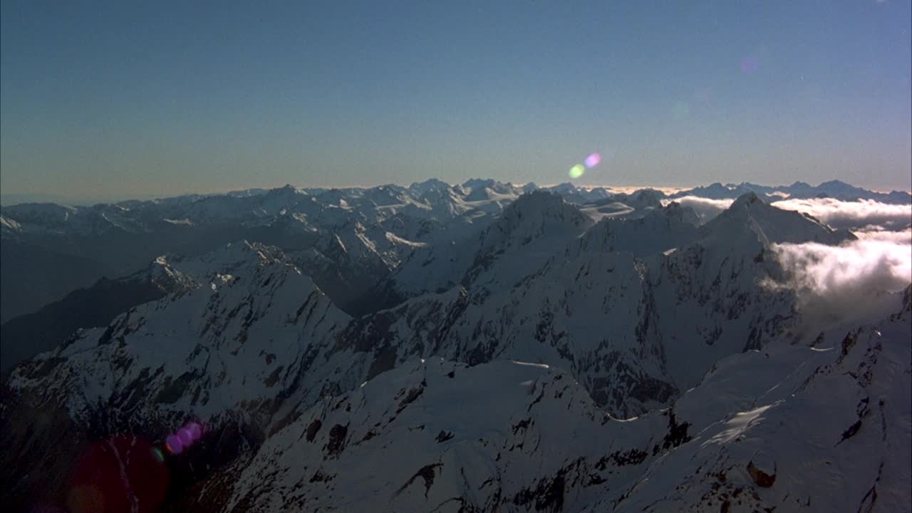 在白雪皑皑的山脉上空飞行。看雪山的山峰。看到镜头耀斑从太阳在右上角和晴朗的蓝天。通过两个被雪覆盖的山峰之间的缺口和下山的山坡倒挂。参见图8中的pov漂移视频素材