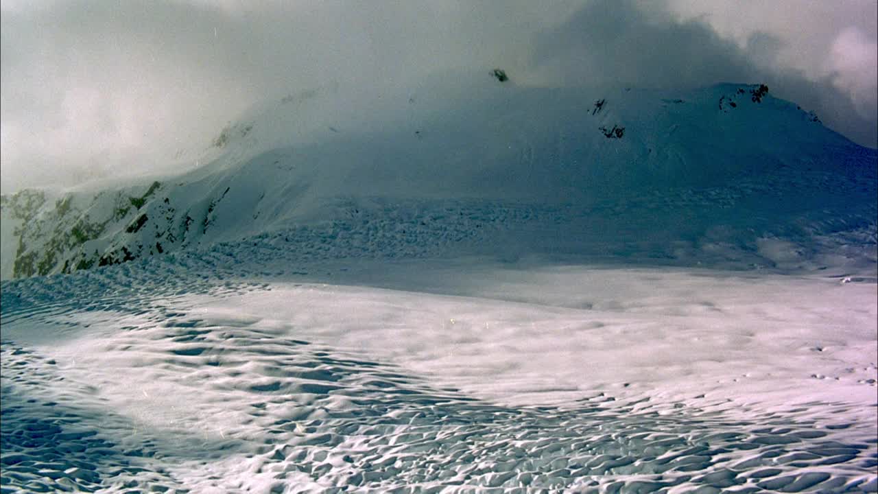 在积雪覆盖的冰川或山坡上的空中飞行。透过浓密的积云，在背景中看到积雪覆盖的山脉和山丘的阴影。视频素材