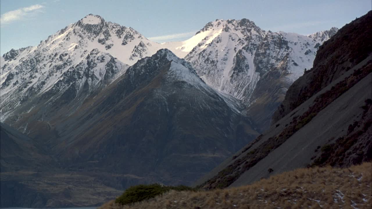 远处背景中雪山山峰的中等角度。山坡前景与两个登山者或徒步旅行者步行下山从右到左。在背景中看到雪山的山谷。视频素材