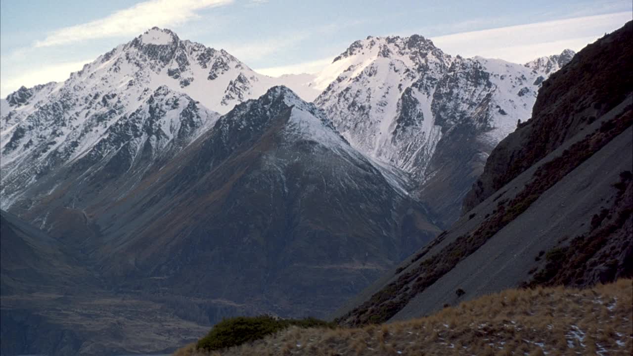 远处背景中雪山山峰的中等角度。山坡前景与两个徒步者或登山者步行下山从右到左。在背景中看到雪山的山谷。视频素材