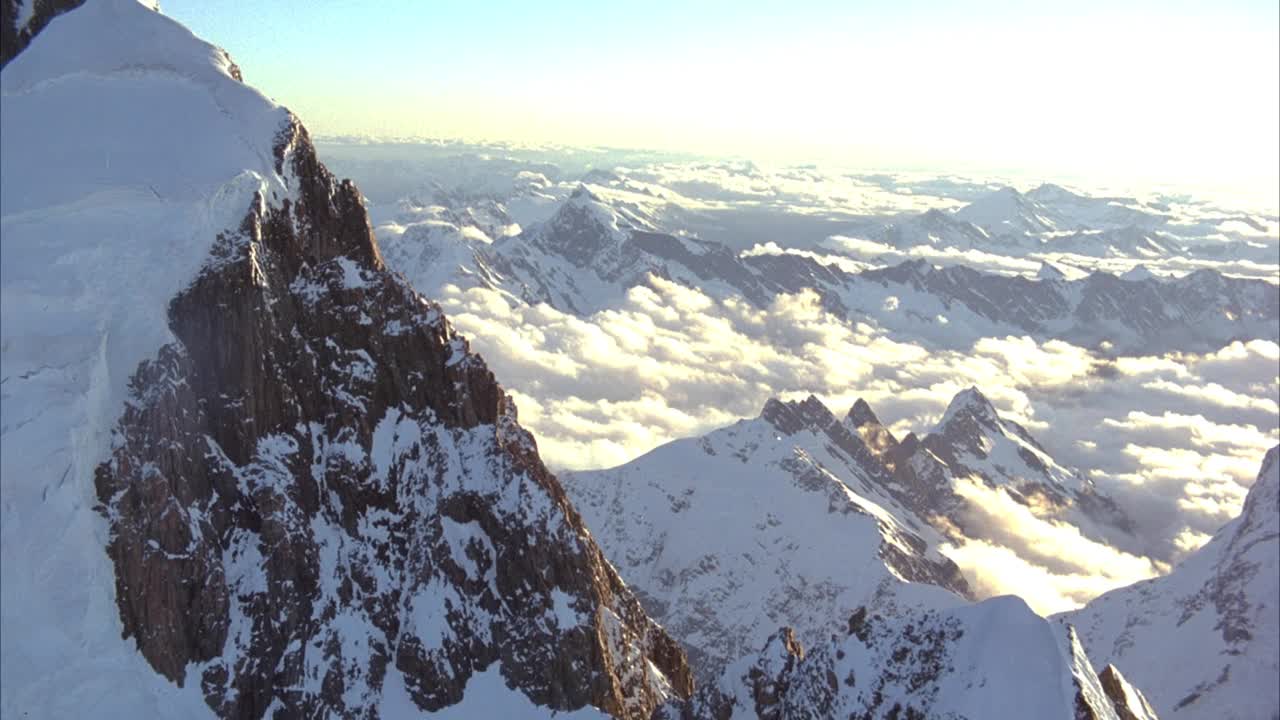 沿白雪皑皑的山脉脊背或山峰的凹地空中飞行。当pov顺时针旋转时，关注arete。拉回至阿雷特侧坡。透过云层可以看到雪山和小山峰。背景是清澈的蓝天。镜头光晕,晕光。视频素材