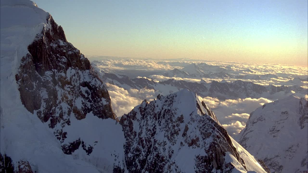 沿白雪皑皑的山脉脊背或山峰的凹地空中飞行。当pov顺时针旋转时，关注arete。拉回至阿雷特侧坡。透过云层可以看到雪山和小山峰。背景是清澈的蓝天。镜头光晕,晕光。视频素材