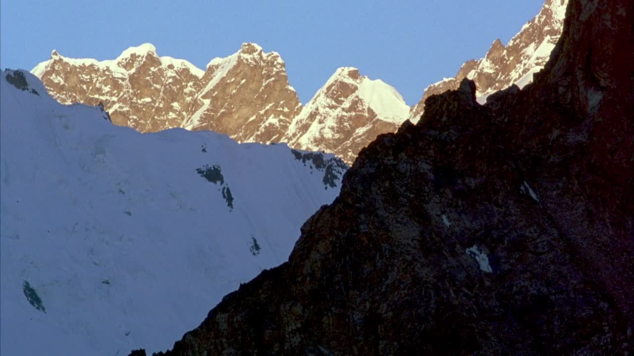 中等角度的岩石灰色的山坡包围在阴影中。看到雪山斜坡和山峰在背景和清澈的蓝天。视频素材