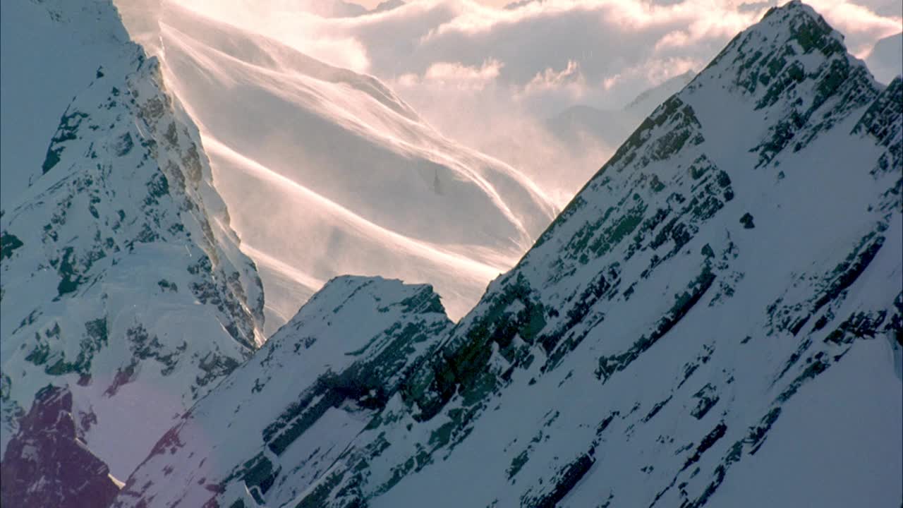 在积雪覆盖的山上空中飞行。看到雾或云沿着雪坡向上滚动。在背景中可以看到低垂的积云挡住了山脉。拉回来看镜头耀斑和光晕来自太阳。视频素材