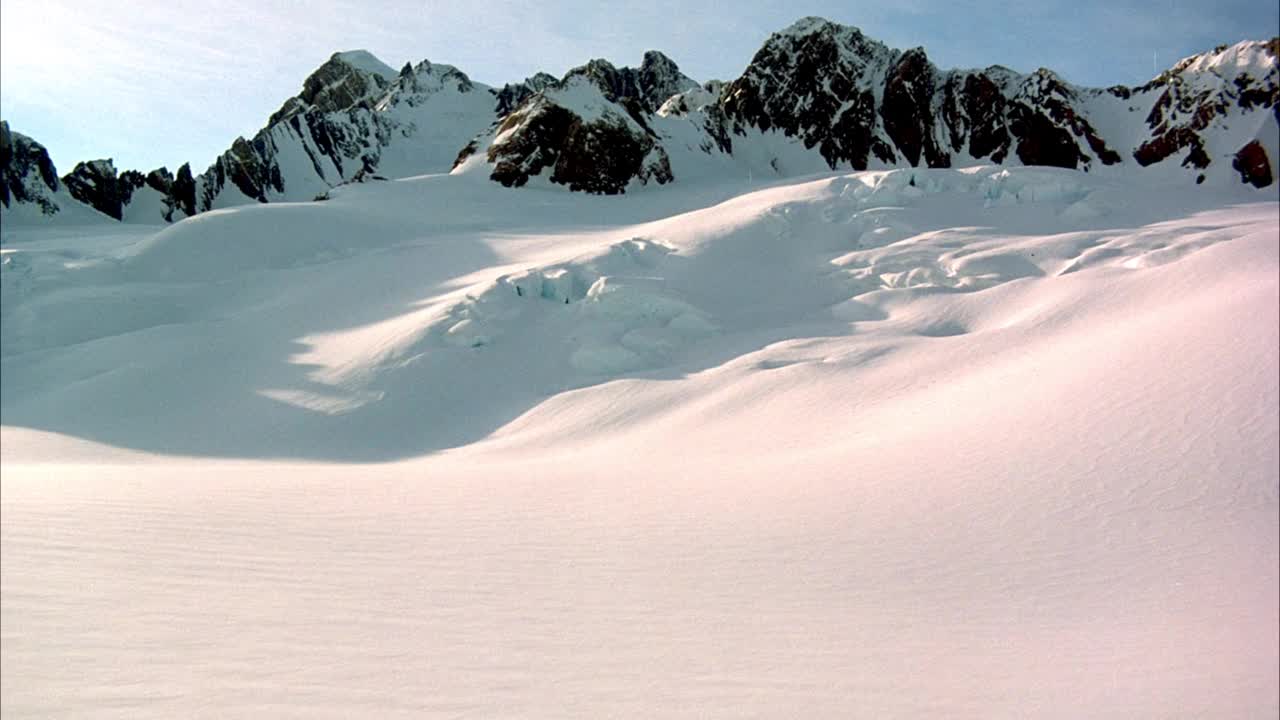 在积雪覆盖的地面和冰川形成的空中拍摄。在背景中看到雪山。看有卷云的蓝天。看镜头从太阳耀斑。视频素材