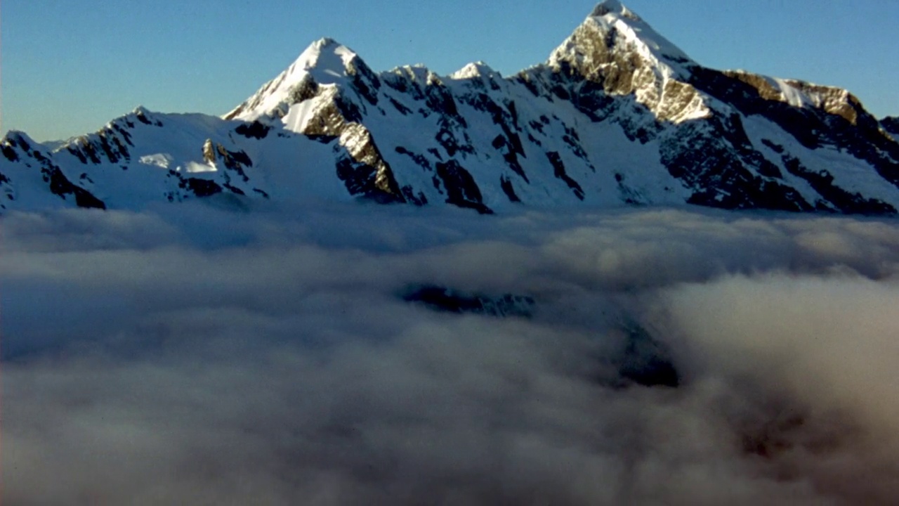 空中的积雪覆盖的山脉和地面附近的云层。在背景中看到清澈的蓝天。相机向各个方向摇摄。视频素材