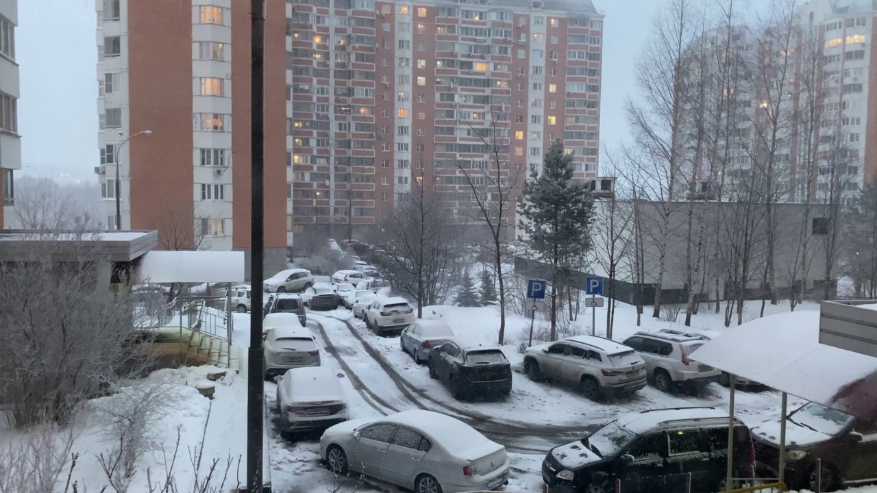 莫斯科高层住宅小区下起了第一场雪。从窗口看出去。冬天。汽车停在入口处。光秃秃的树，路灯，房子附近的操场。4 k视频素材