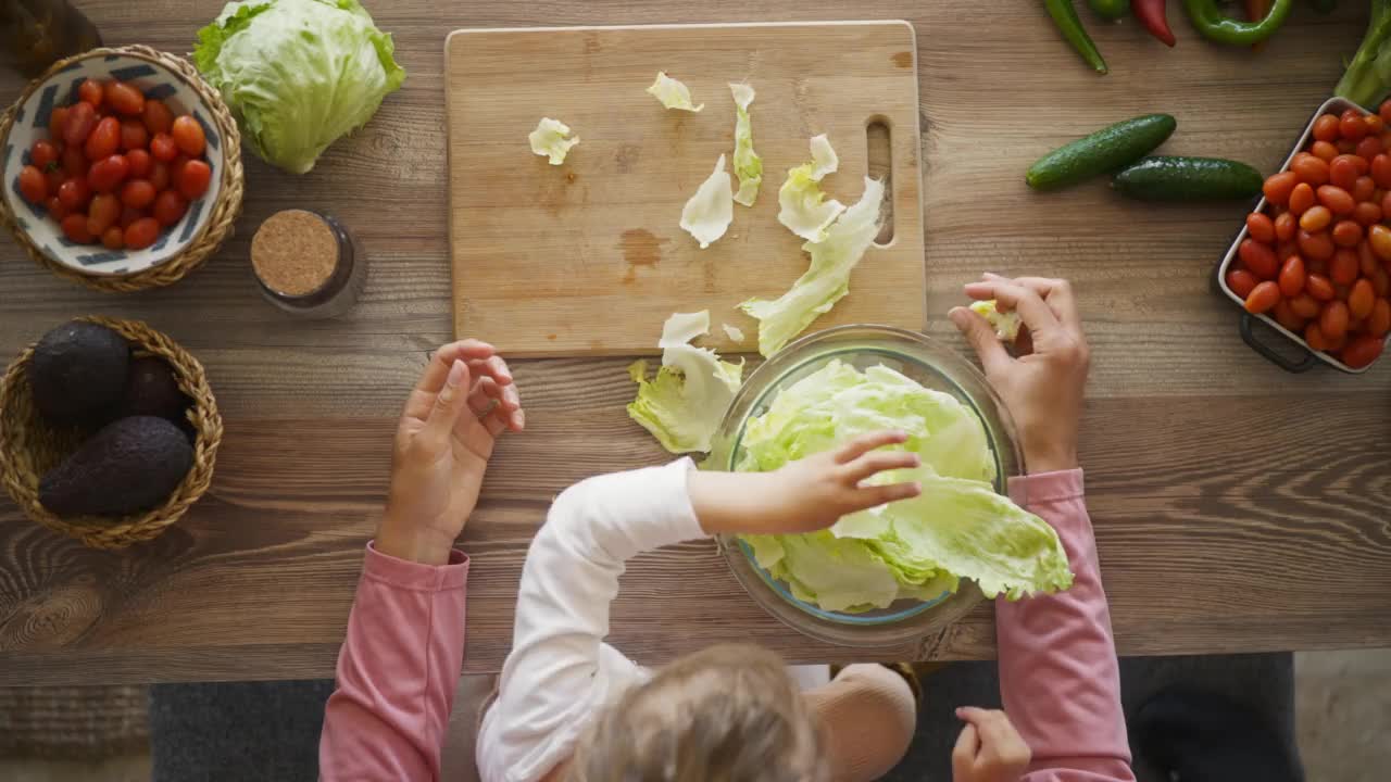 一个女人带着她的女儿在厨房里准备沙拉。女孩和她妈妈一起做饭。女孩片蔬菜。视频素材