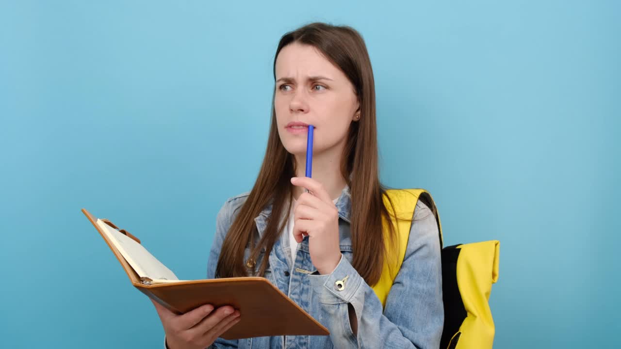 肖像恼怒的女孩十几岁的学生穿牛仔夹克和黄色背包写在练习本笔记，在工作室的蓝色背景上孤立地摆姿势。大学教育视频素材