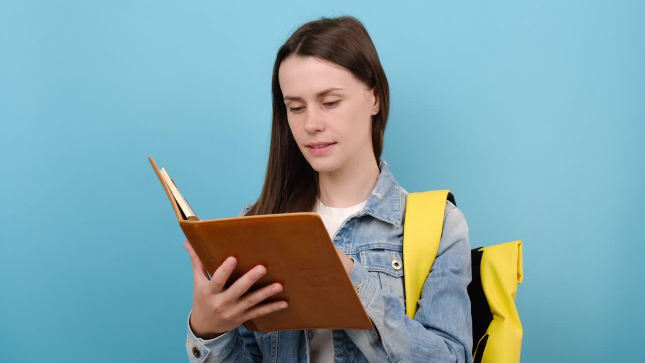 肖像女孩十几岁的学生穿牛仔夹克和黄色背包读书想梦，在工作室的蓝色背景墙孤立地摆姿势。高中、大学、大学教育理念视频素材