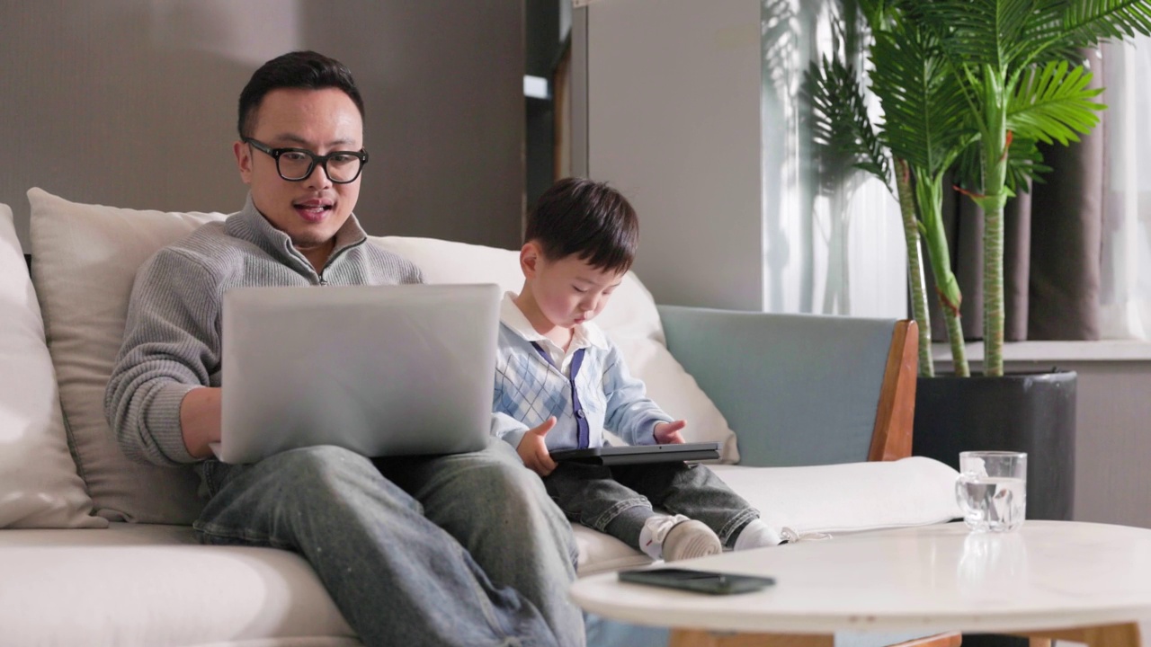 中年男子在沙发上工作，他的儿子在他旁边玩视频素材画面1