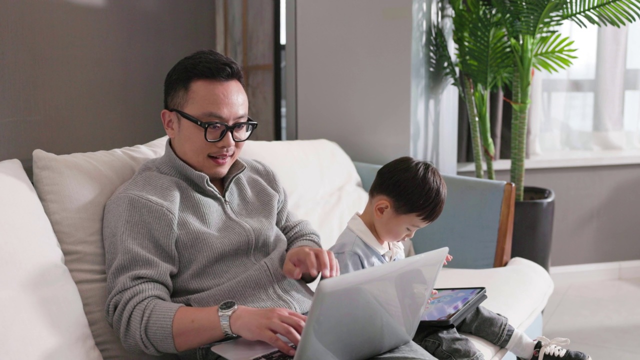 父亲在沙发上工作，儿子在他旁边玩视频素材画面3