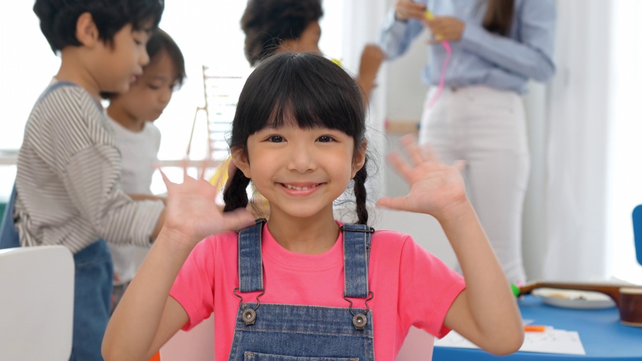 快乐的小女孩微笑着和她摊手的另一个朋友背景在教室里，幼儿园教育学校。多民族幼儿教师和学生的课堂。幼儿园的概念。视频素材