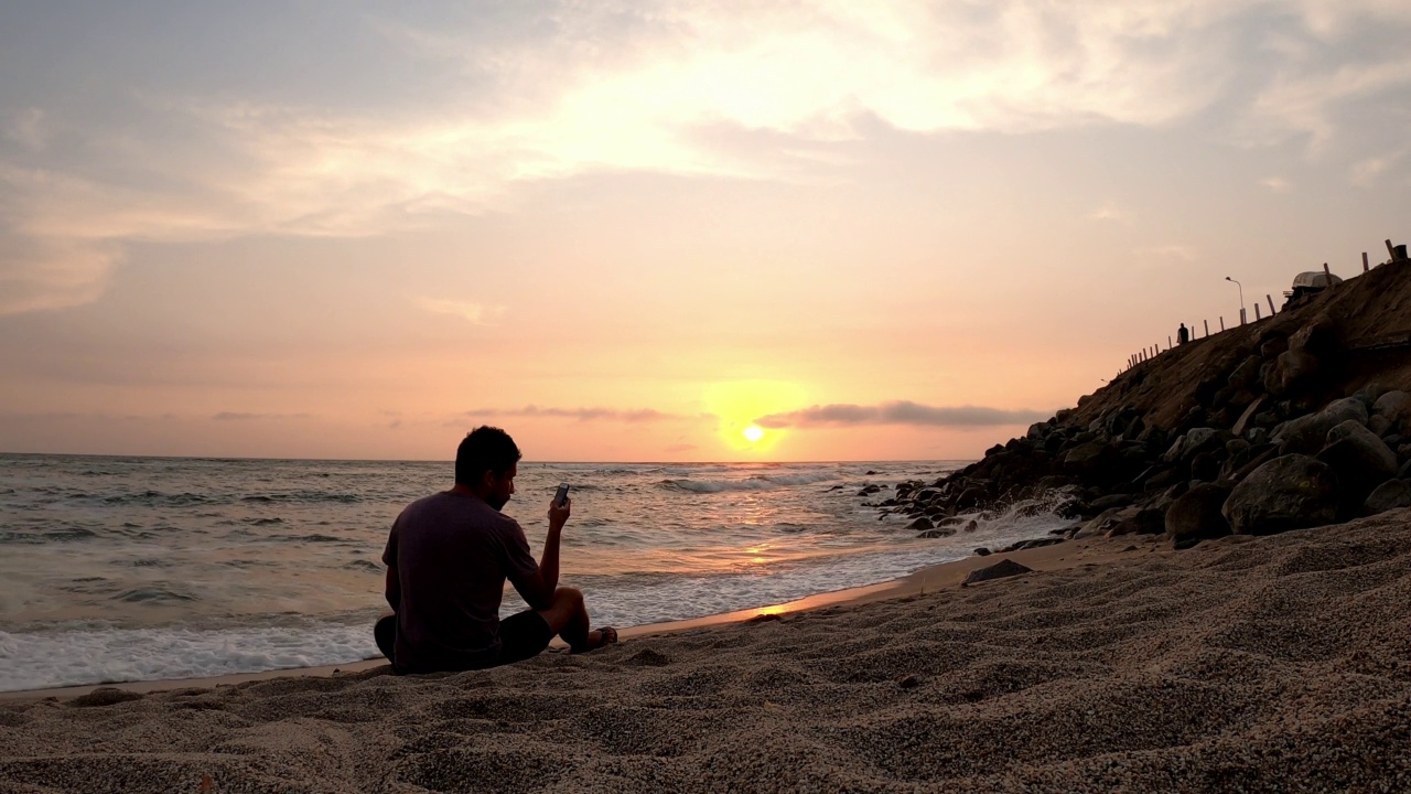 一个男人坐在沙滩上看手机，4k的海景是沙滩上的日落和地平线上的太阳视频素材