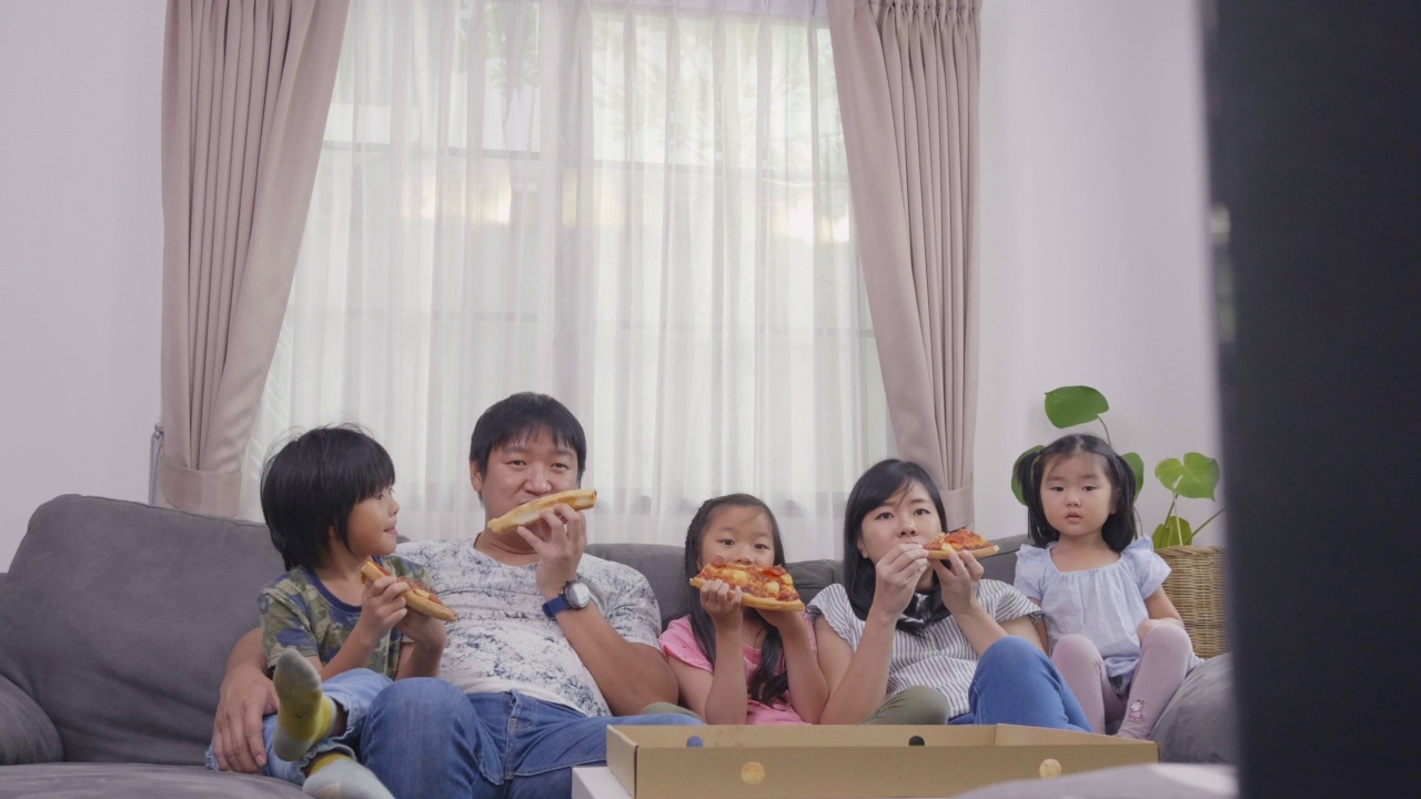 幸福的亚洲家庭喜欢在假日在家的沙发上吃披萨和看电视视频素材画面3