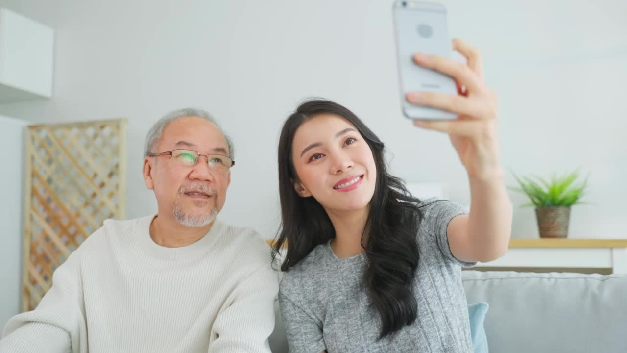 亚洲可爱的家庭，女儿用电话与资深父亲视频通话。年轻漂亮的女孩和年长的大龄男性坐在沙发上享受通过互联网交流，在家里的客厅一起聊天视频素材