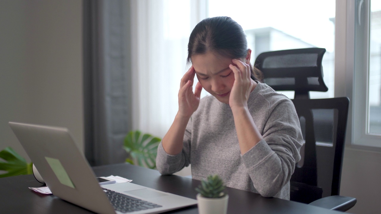 一名亚洲女性在家用笔记本电脑办公时头痛。视频素材