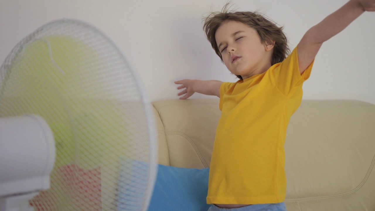 热天站在沙发上的男孩。炎炎夏日，孩子在家用电风扇吹凉风。忍受高温在通风柜前用电子冷却器冷却自己视频素材