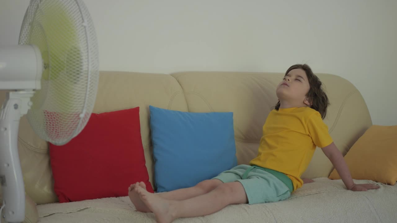 热天坐在沙发上的男孩。炎炎夏日，孩子在家用电风扇吹凉风。忍受高温在通风柜前用电风扇冷却器冷却自己视频素材
