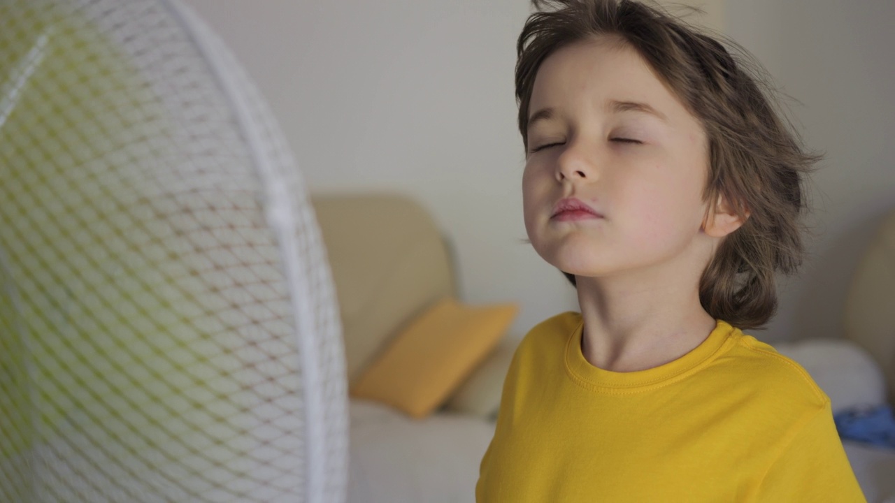 炎炎夏日，孩子在家用电风扇吹凉风。忍受高温在通风柜前用电风扇冷却器冷却自己。站在范面前的男孩。视频素材
