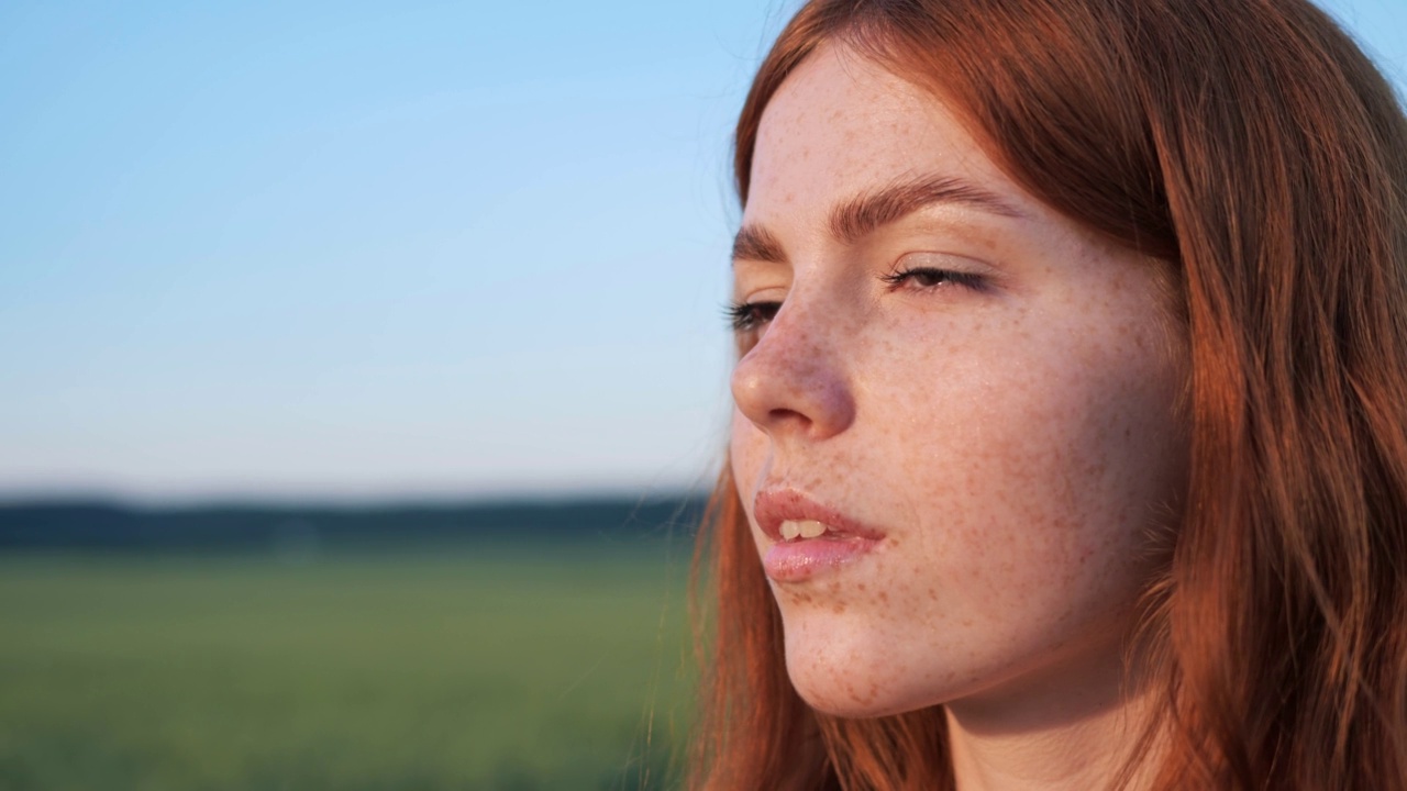 美丽的女性姜黄脸与雀斑的自然在日落。女人向前看，有长长的红头发，美丽的孩子的脸。有一双棕色眼睛的迷人女孩站在户外的田野里。视频素材