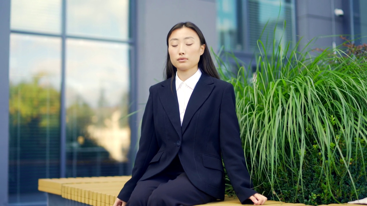 亚洲女性办公室职员放松，冥想户外坐在城市背景的长椅上，城市公园休息休息商务女性员工在正式的西装，闭上眼睛视频素材