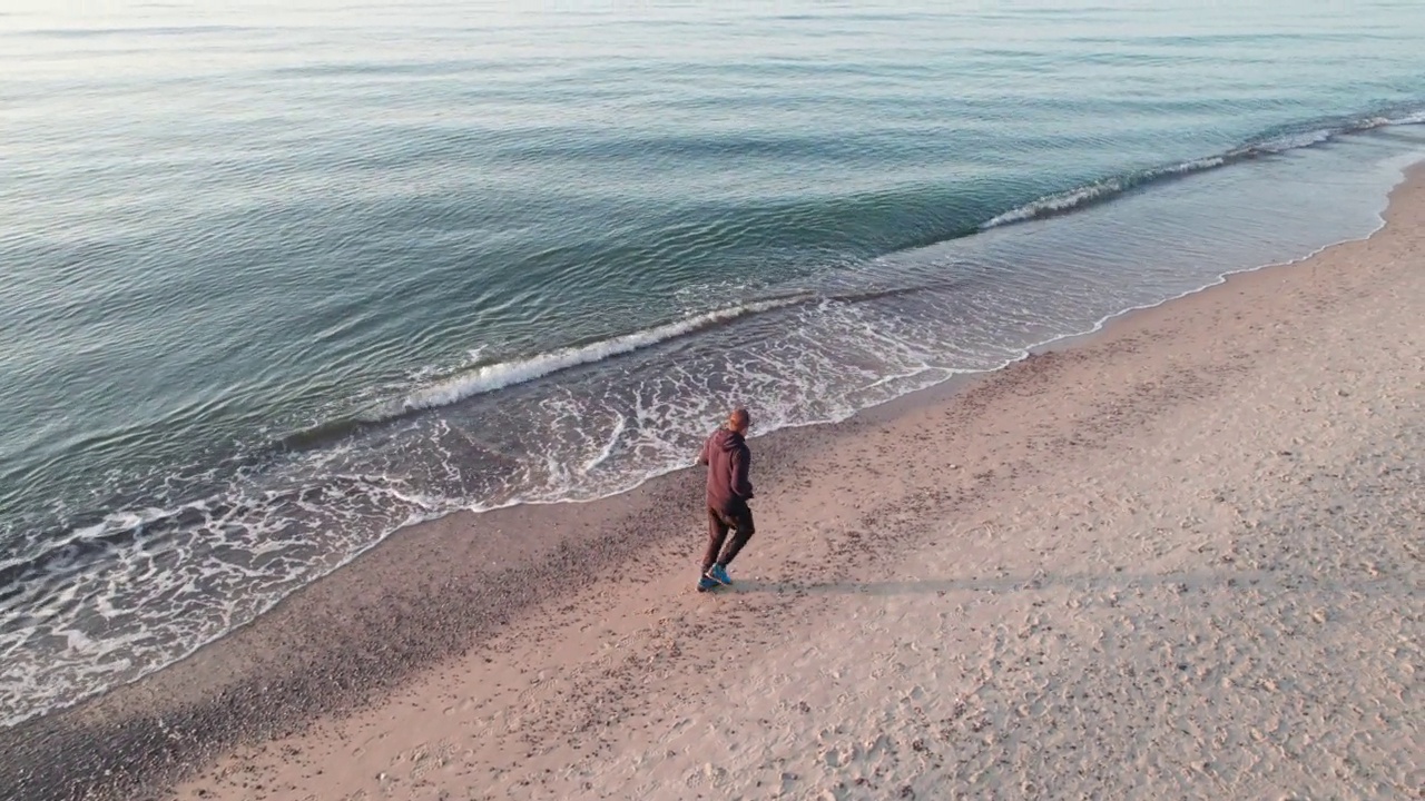 一个跑步者，在日落的时候，他沿着海滩沿着波浪跑。视频素材