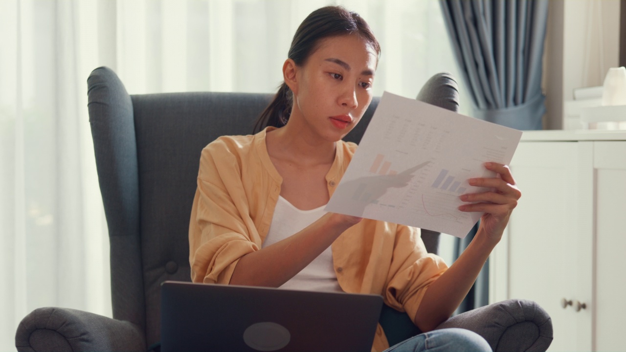 年轻迷人的亚洲女女孩或大学生坐在沙发椅上用电脑笔记本电脑在家里客厅的报告工作文书检查数据图表。视频素材