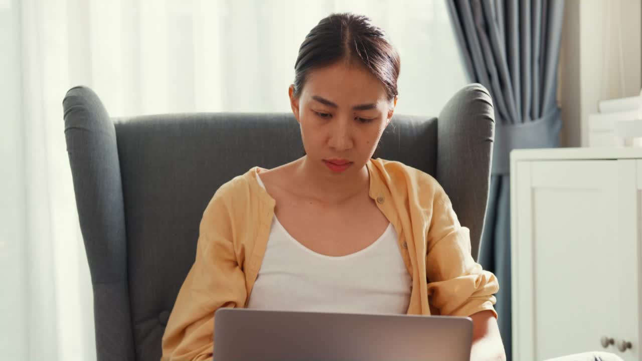 年轻的亚洲女学生或大学生坐在沙发椅上拿着电脑笔记本电脑感到疼痛，在家里的客厅里因加班劳累而伤了肩膀。视频素材