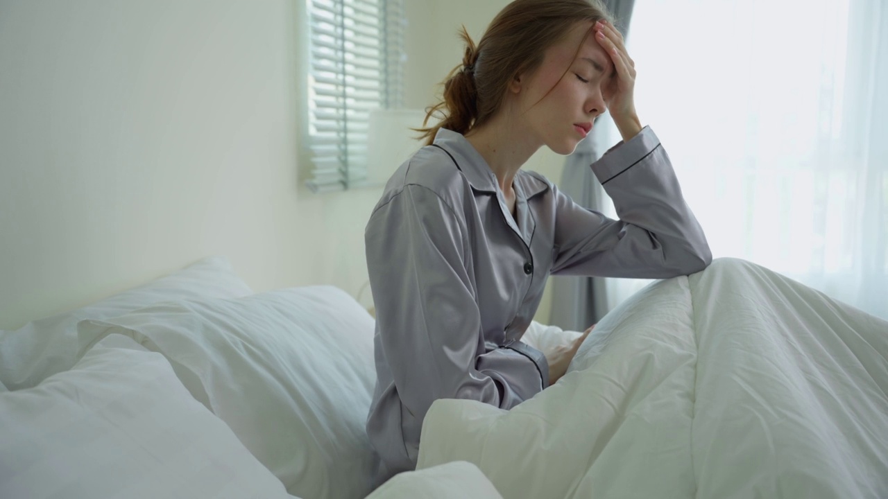 白人年轻女孩穿着睡衣坐在卧室的床上感到恶心。迷人的美丽女子摸着额头，感觉不好发烧和头痛后，早上醒来在家里的床上。视频素材