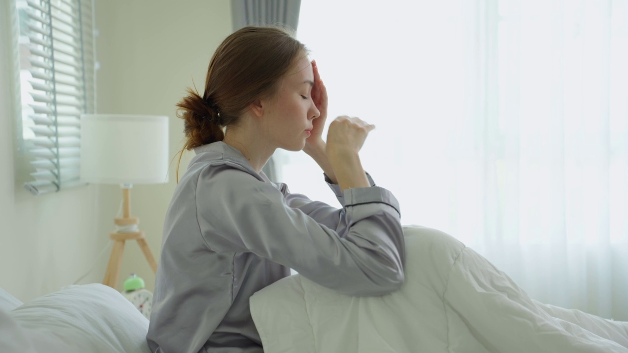 白人年轻女孩穿着睡衣坐在卧室的床上感到恶心。迷人的美丽女子摸着额头，感觉不好发烧和头痛后，早上醒来在家里的床上。视频素材