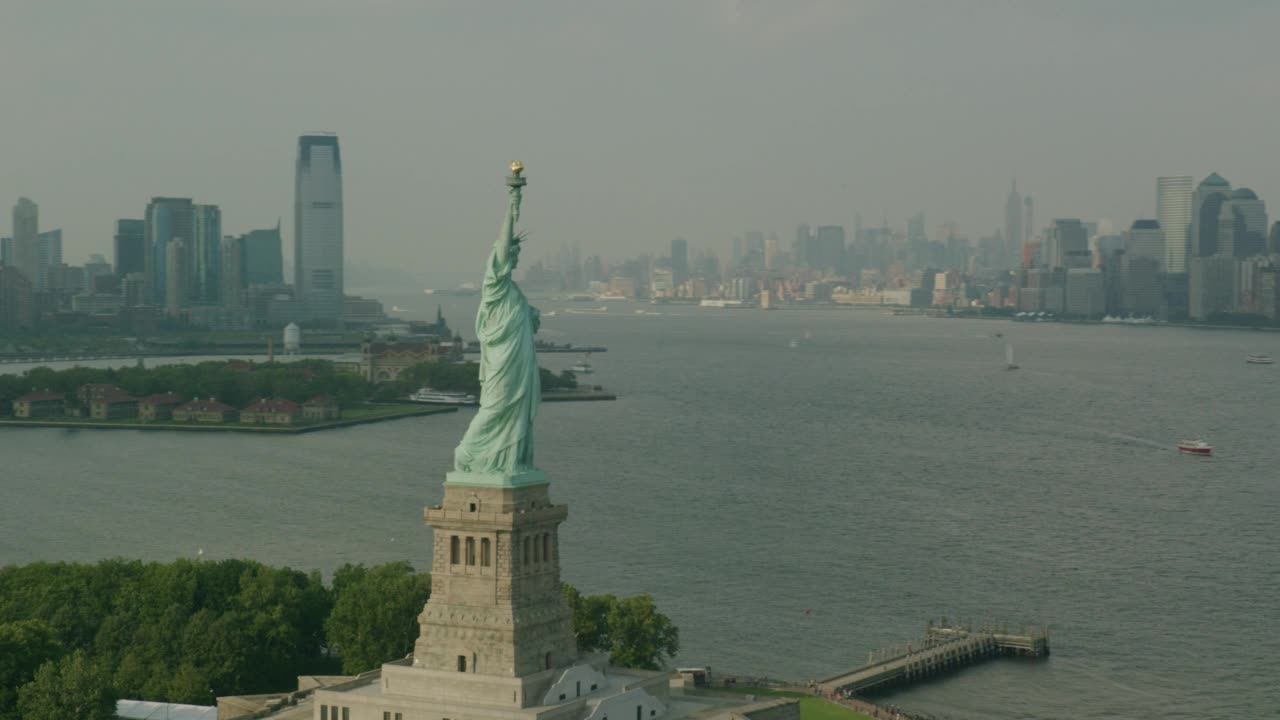 自由女神像和自由岛的空中全景图。埃利斯岛和渡轮可见。镜头摇摄到纽约天际线的更广视野，一个世界贸易中心或自由塔和帝国大厦可见。纽约港和哈德逊河之间视频素材