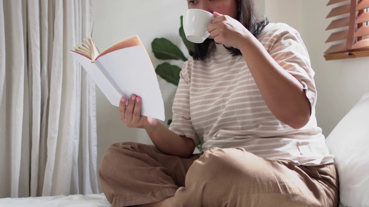 快乐的年轻女子坐在床上喜欢看书，拿着一杯咖啡或茶在家里放松早上视频素材