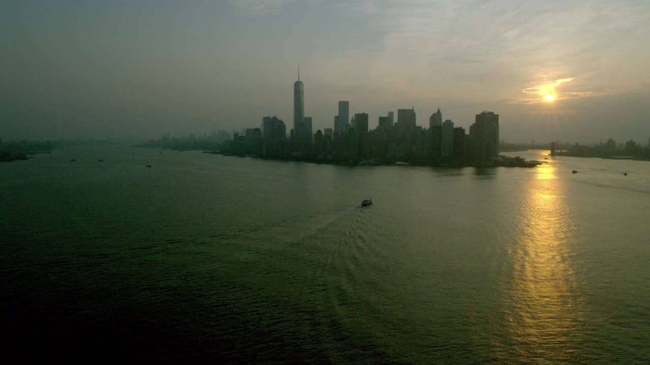 航拍渡船在纽约港从左向右驶向纽约市。哈德逊河，东河，布鲁克林大桥，炮台公园，和一个世界贸易中心或自由塔可见。可能是日出还是日落视频素材