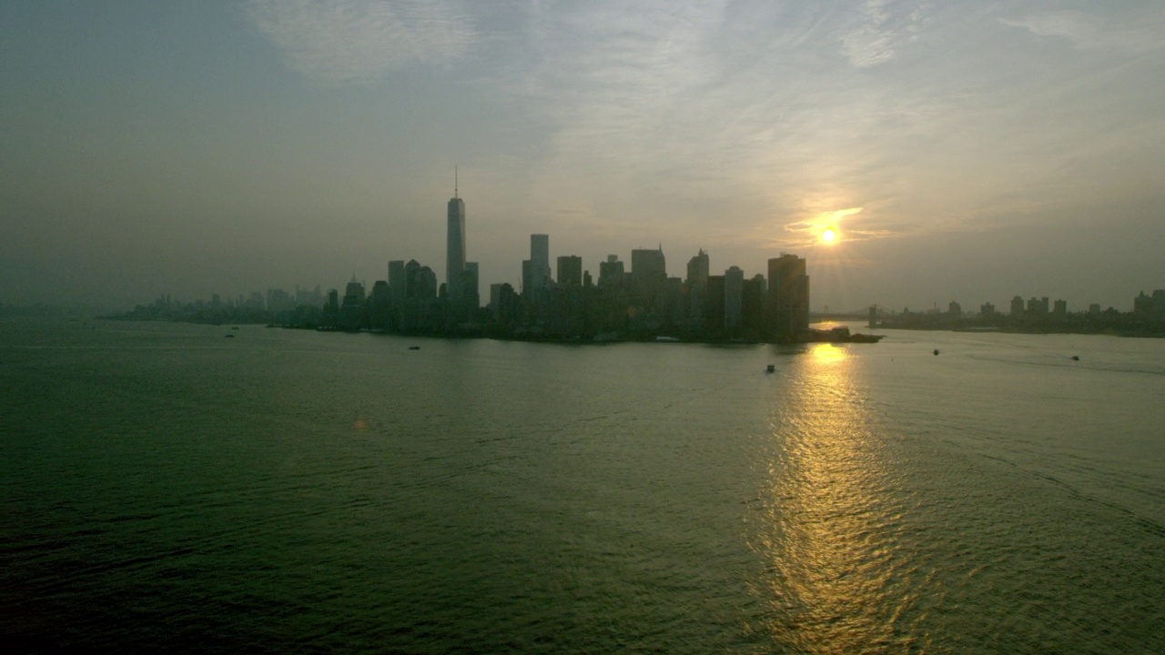 从右到左的空中移动渡轮在纽约港向纽约市前进。哈德逊河，东河，布鲁克林大桥，炮台公园，和一个世界贸易中心或自由塔可见。可能是日出还是日落视频素材