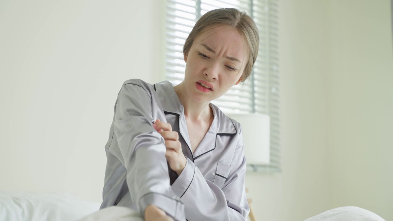 年轻的白人妇女在早上醒来后抓挠手。美丽迷人的女孩躺在床上的卧室遭受痒的手臂皮肤过敏反应，昆虫叮咬，皮炎。视频素材