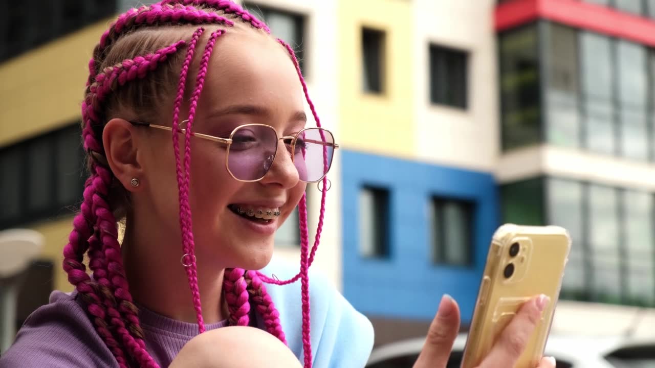 一个带着粉色辫子的微笑的白人年轻时髦女孩在街道背景上使用智能手机的肖像。夏天的概念。Z一代的风格。社交媒体的概念视频素材画面1