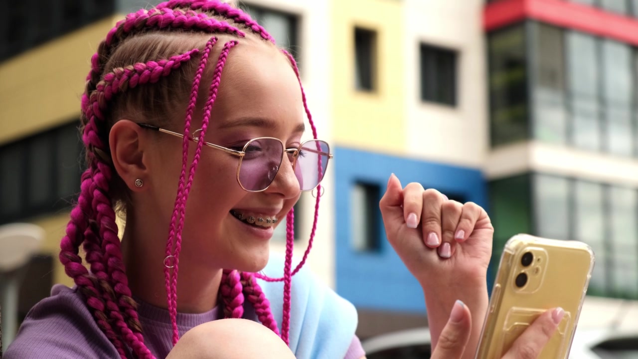 一个带着粉色辫子的微笑的白人年轻时髦女孩在街道背景上使用智能手机的肖像。夏天的概念。Z一代的风格。社交媒体的概念视频素材画面2