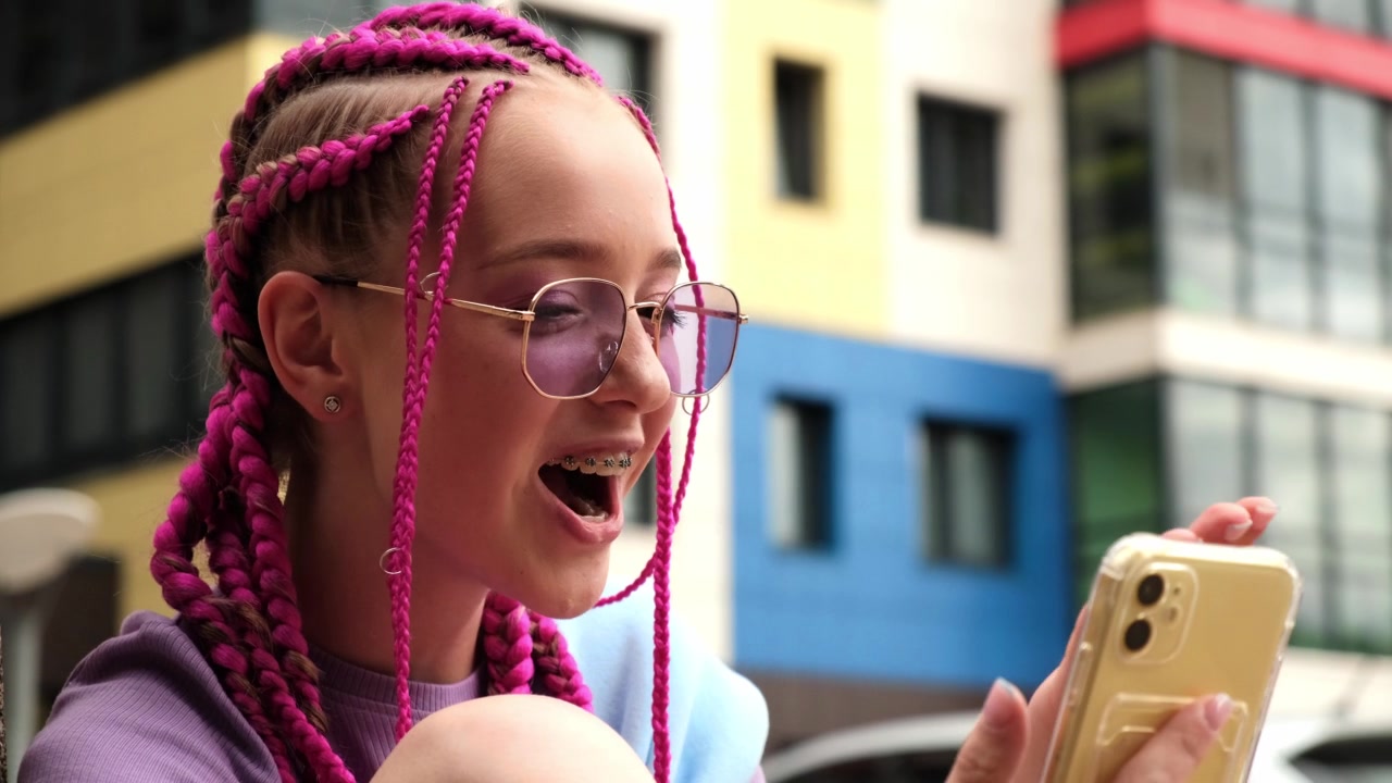 一个带着粉色辫子的微笑的白人年轻时髦女孩在街道背景上使用智能手机的肖像。夏天的概念。Z一代的风格。社交媒体的概念视频素材画面4