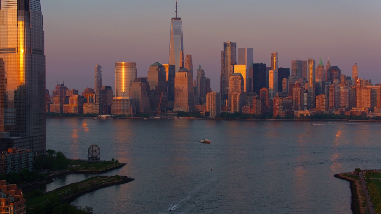 日落时分，从泽西城的保卢斯胡克(Paulus Hook)可以看到远处的哈德逊河(Hudson River)上的世界贸易中心(One World Trade Center)和沿河航行的船只。带有向后上升的摄像机运动的航拍视频。视频素材