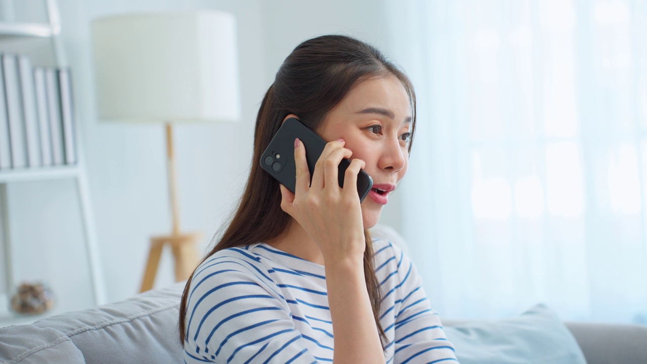 亚洲美女坐在沙发上，在家里打电话。快乐迷人的年轻女孩在家里度过闲暇时光，感到放松，享受在客厅用智能手机交流和讨论。视频素材