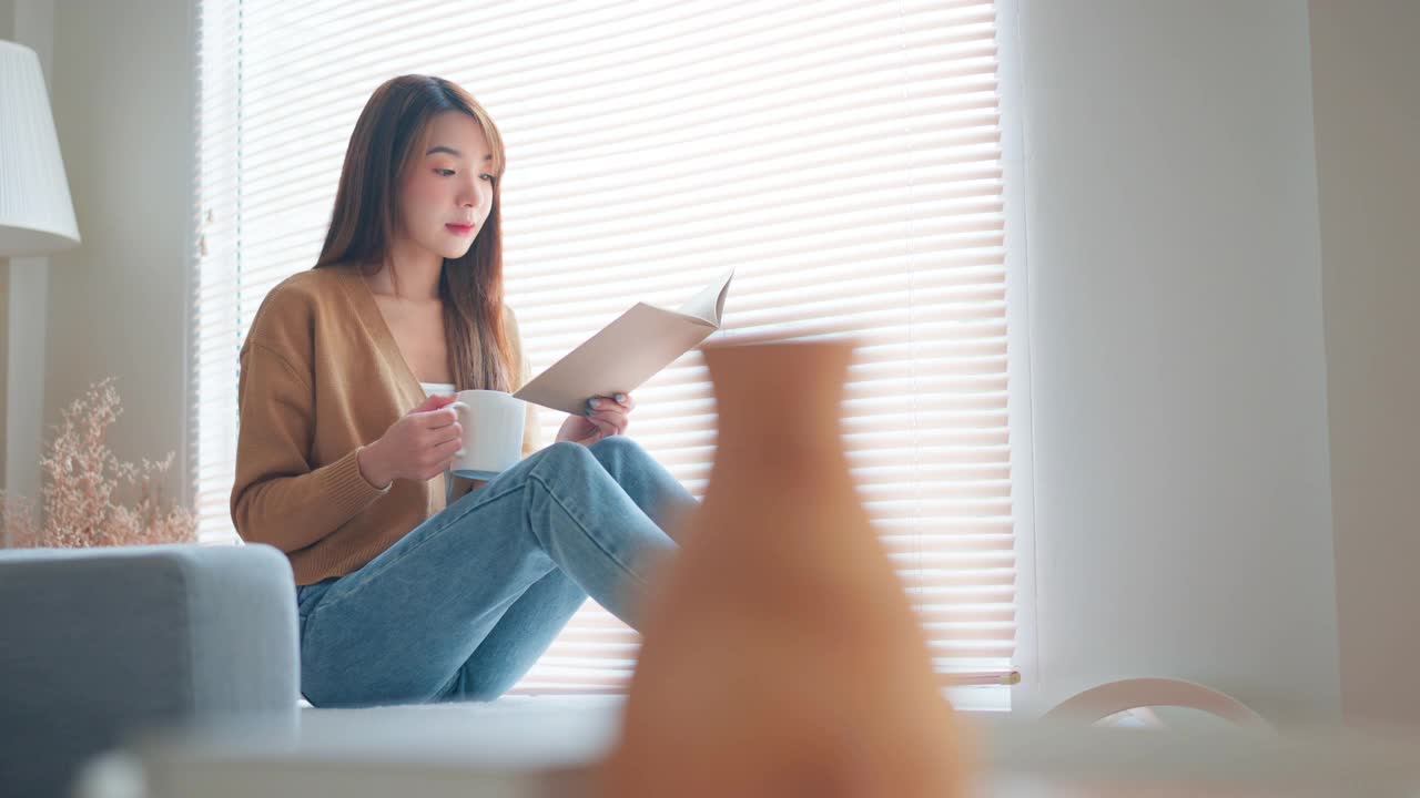 快乐的亚洲女人喝咖啡或茶，在家里的客厅窗前看书放松，生活方式的家的概念。视频素材