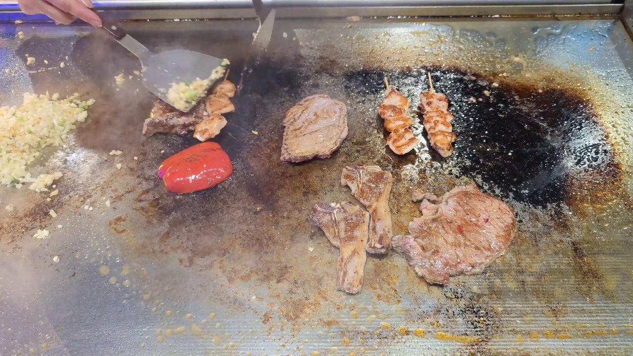 Tenderloin, steak, chicken skewers, peppers and lamb chops are topped with sliced ​​garlic and spring onions by an unrecognizable chef and then fried on a teriyaki grill plate in a restaurant视频素材画面1