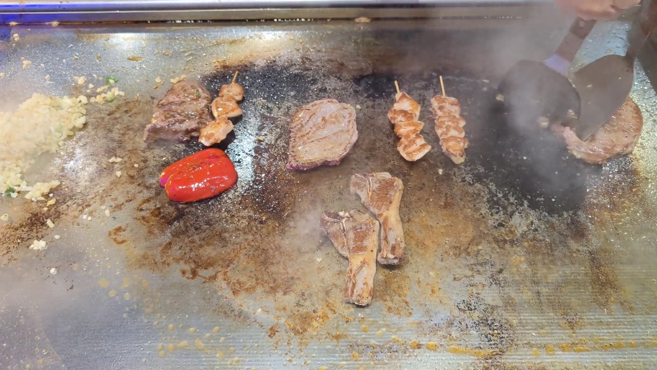 Tenderloin, steak, chicken skewers, peppers and lamb chops are topped with sliced ​​garlic and spring onions by an unrecognizable chef and then fried on a teriyaki grill plate in a restaurant视频素材画面2